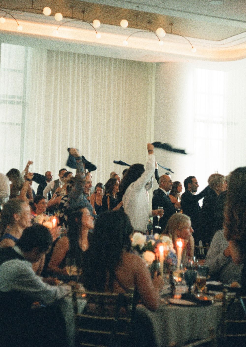 Guests celebrate at a wedding reception dinner, raising their hands in cheer around candlelit tables in an elegant ballroom.