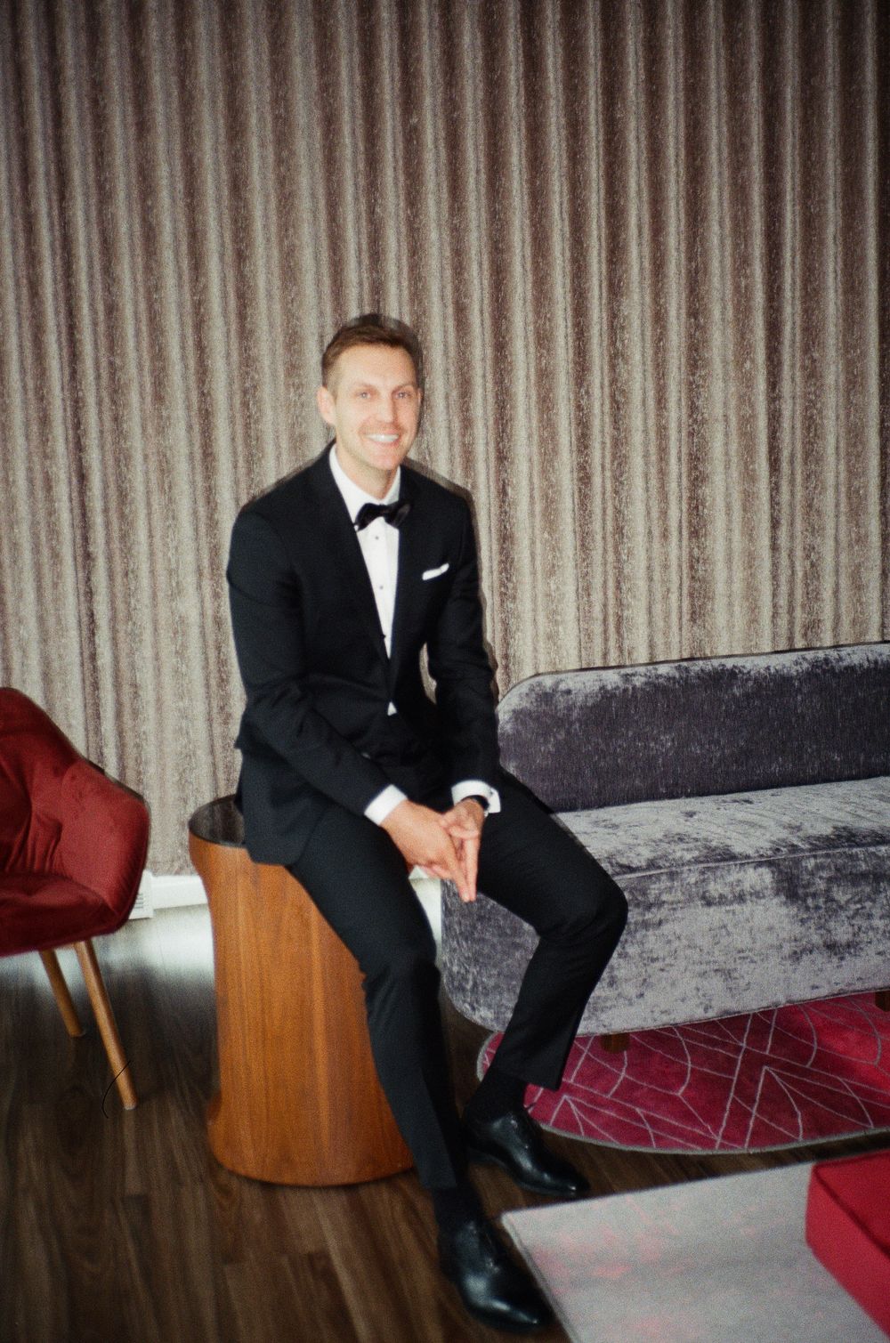 Man in black tuxedo with bow tie sitting on a velvet sofa, smiling in an elegant room.