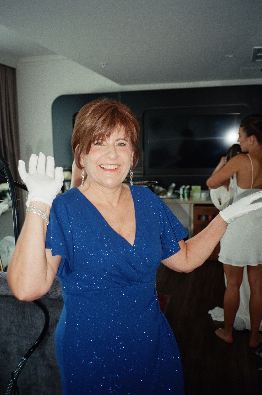 Smiling woman in blue sparkly dress wearing white gloves, waving hands in indoor setting.