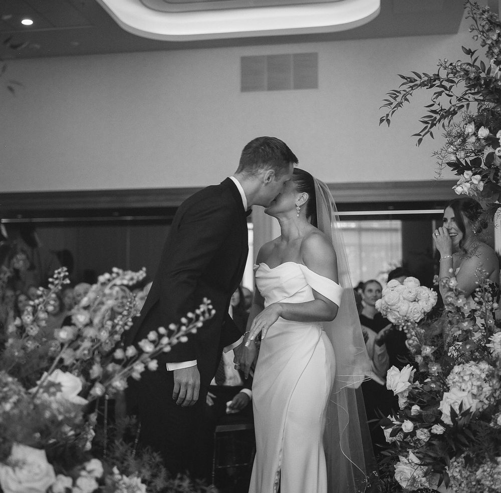 Black and white photo of a bride and groom sharing their first kiss at the altar surrounded by floral arrangements.