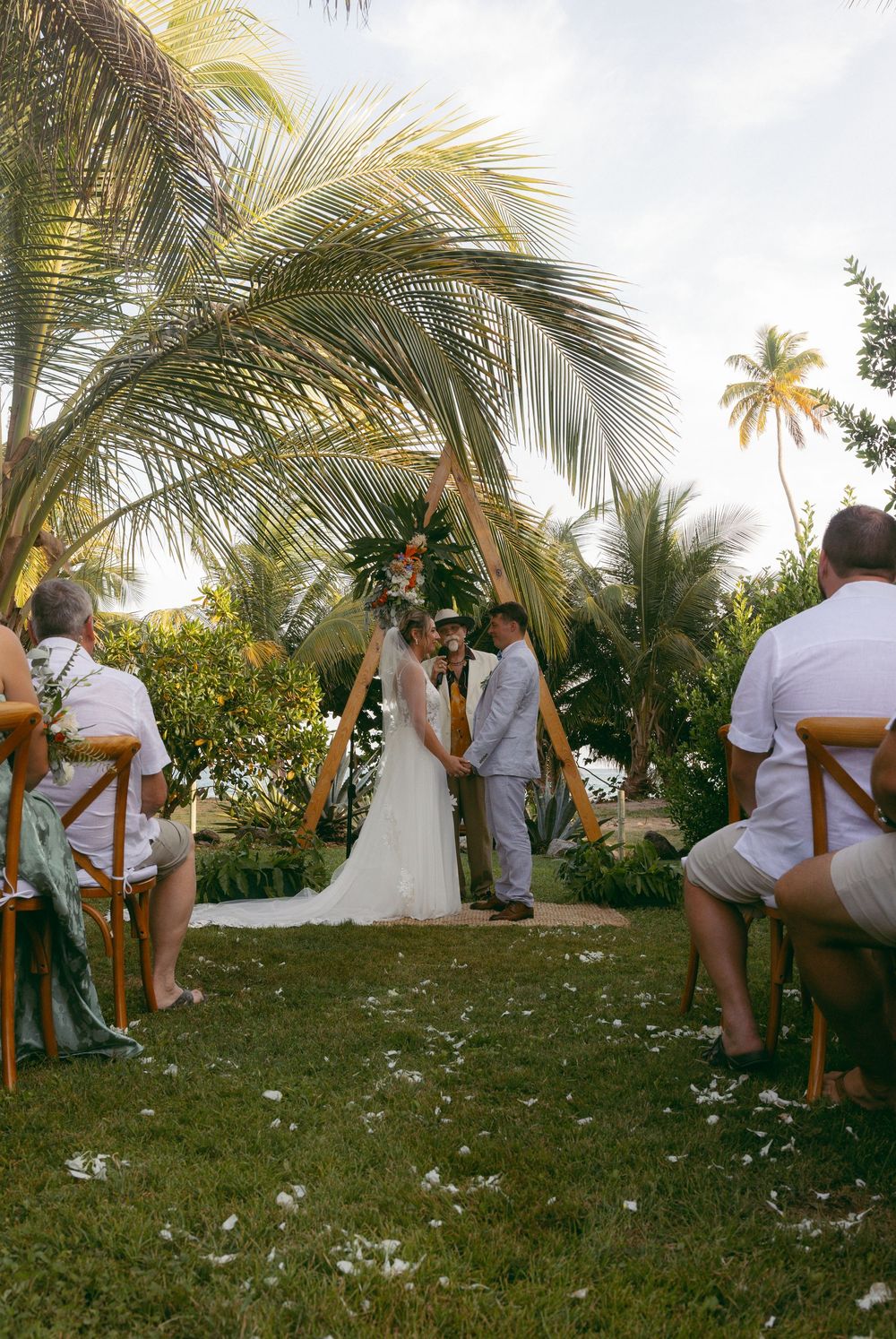Couple exchanging vows at tropical outdoor wedding ceremony under palm trees with floral arch and petal-lined aisle.
