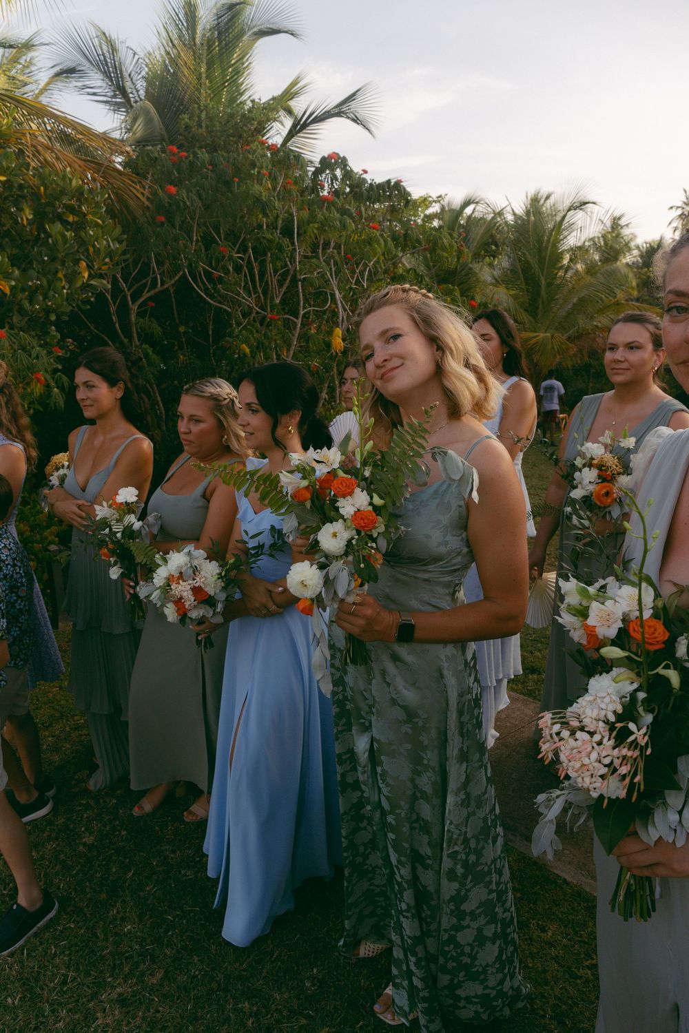 Bridesmaids in blue and green dresses holding floral bouquets at an outdoor tropical wedding ceremony.