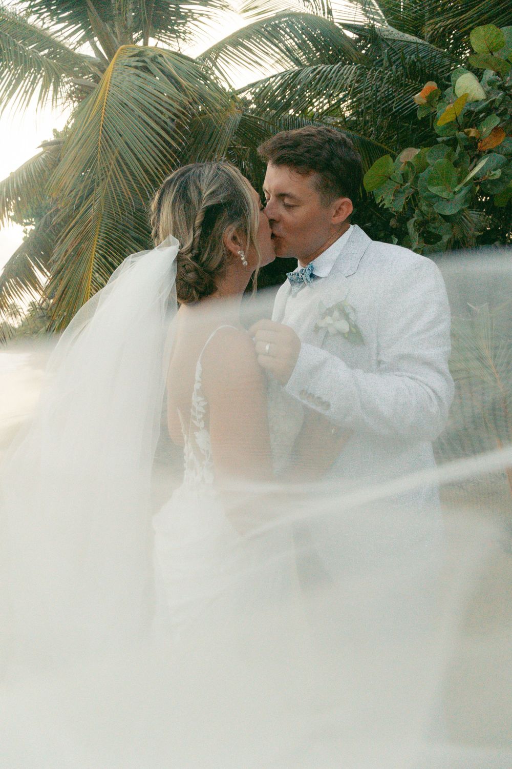 Bride and groom sharing a romantic kiss under palm trees, with a flowing veil, at a tropical beach wedding.