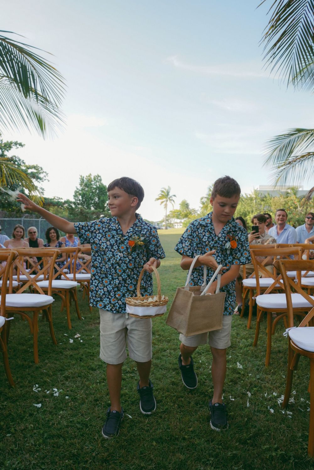Two boys in matching floral shirts scatter flower petals down an outdoor wedding aisle lined with wooden chairs and palm trees.