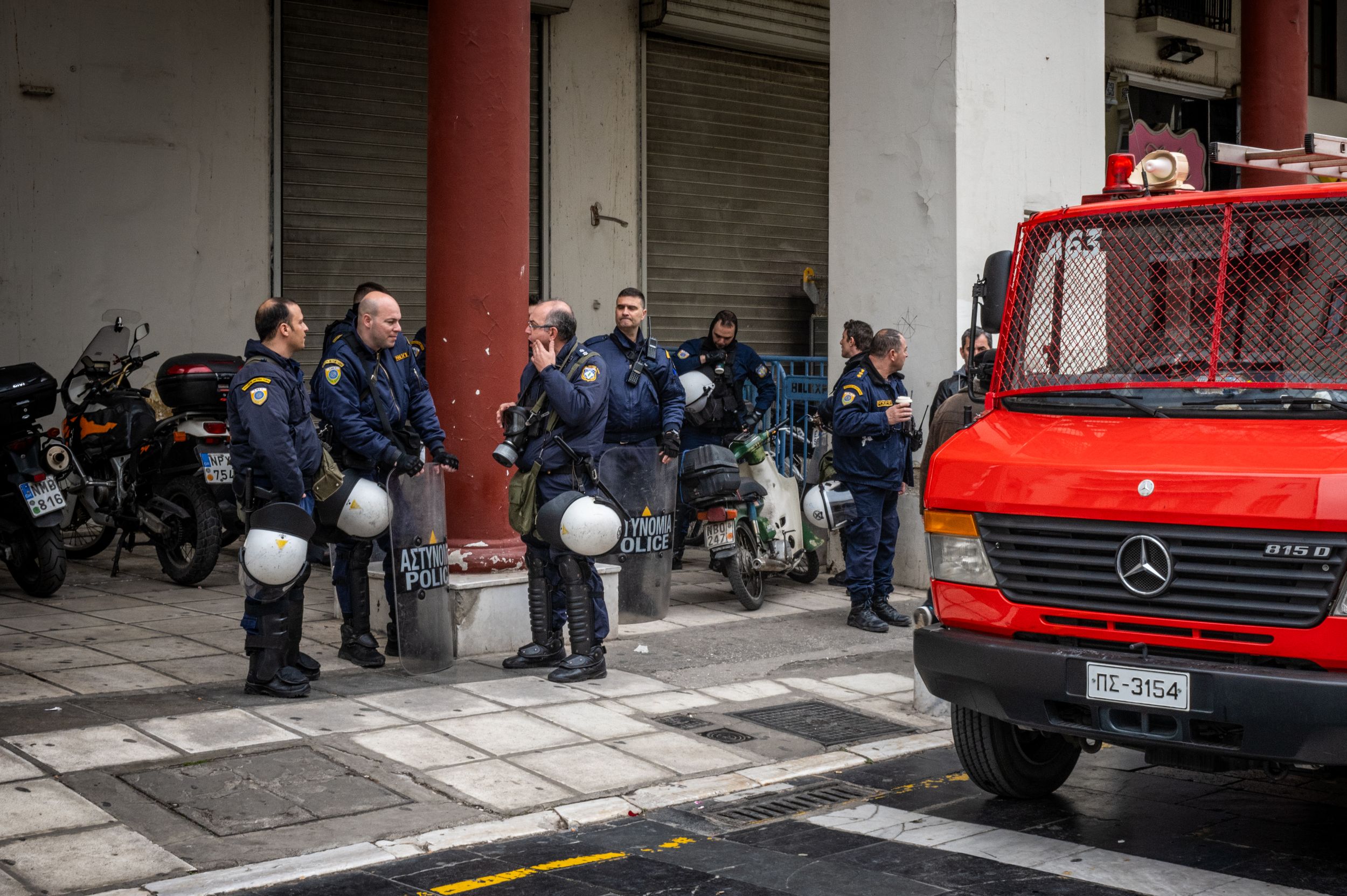 Riot police officers stand near a red Mercedes fire truck on a city street during an emergency response operation.
