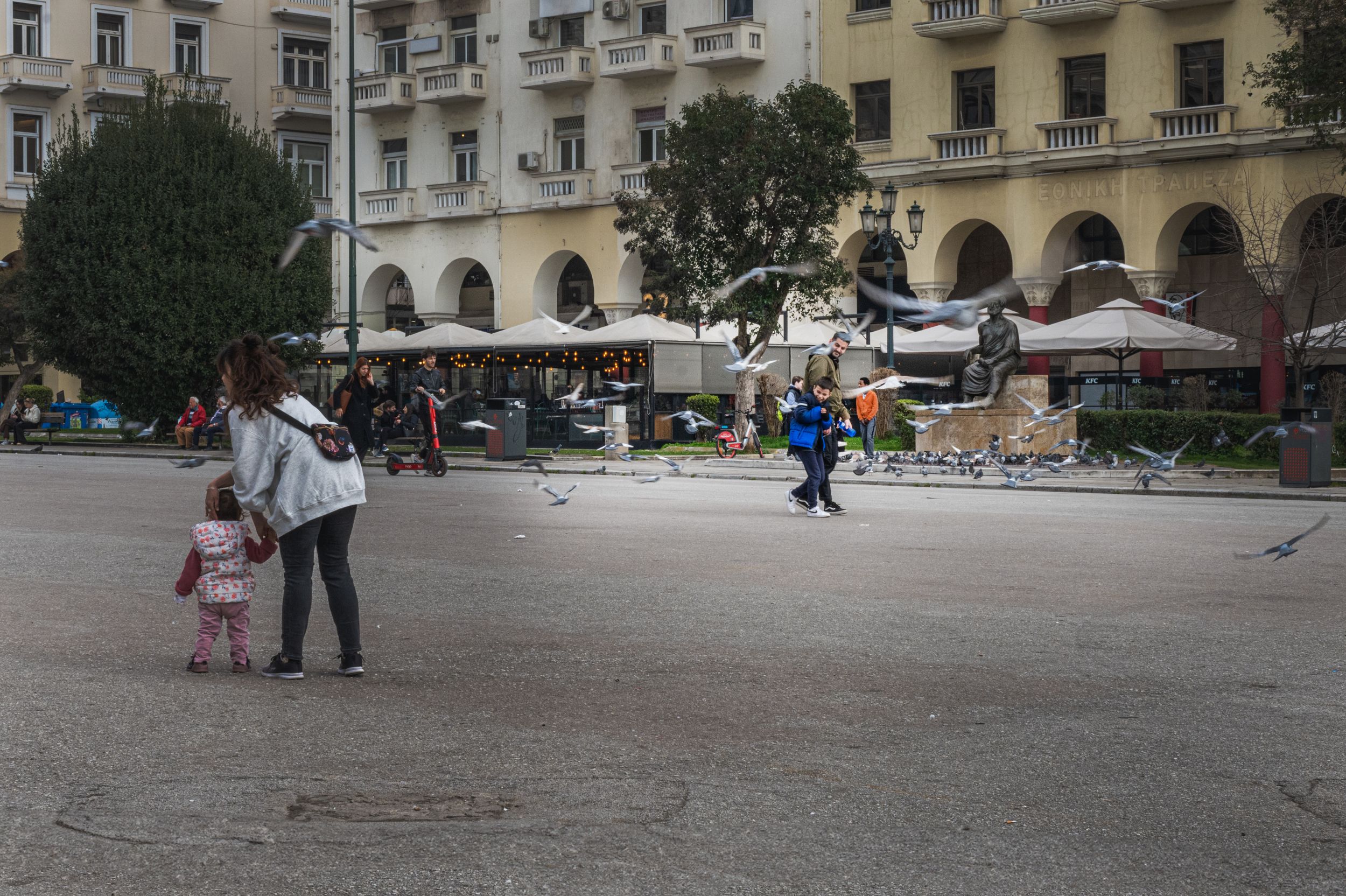 A woman and child stand in a large European city square with outdoor cafes and neoclassical buildings in the background.