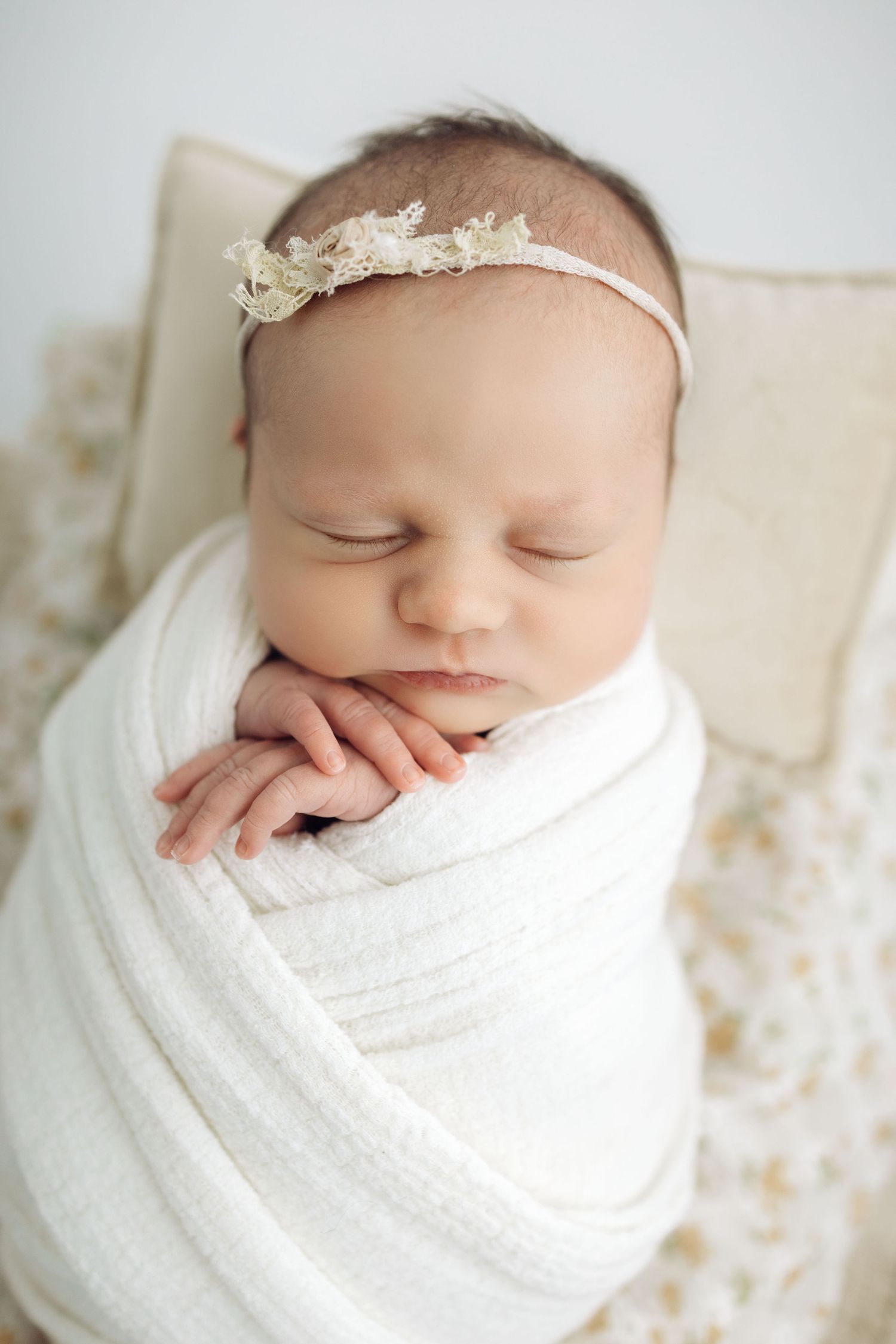 Sleeping newborn baby girl swaddled in white wrap with lace headband, peaceful portrait photography.