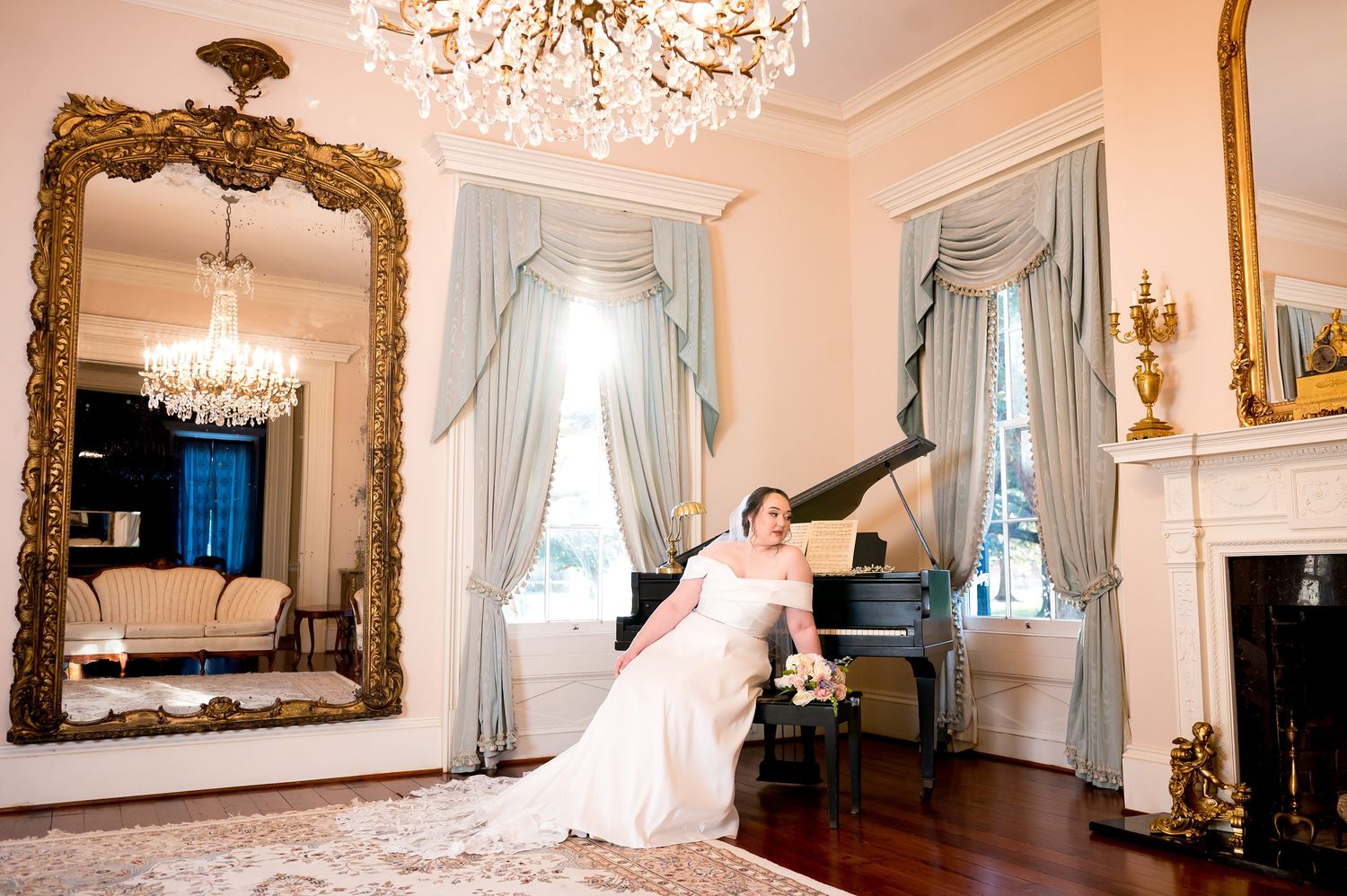 Bride in white gown poses by grand piano in elegant ballroom with chandelier, ornate mirror, and fireplace.