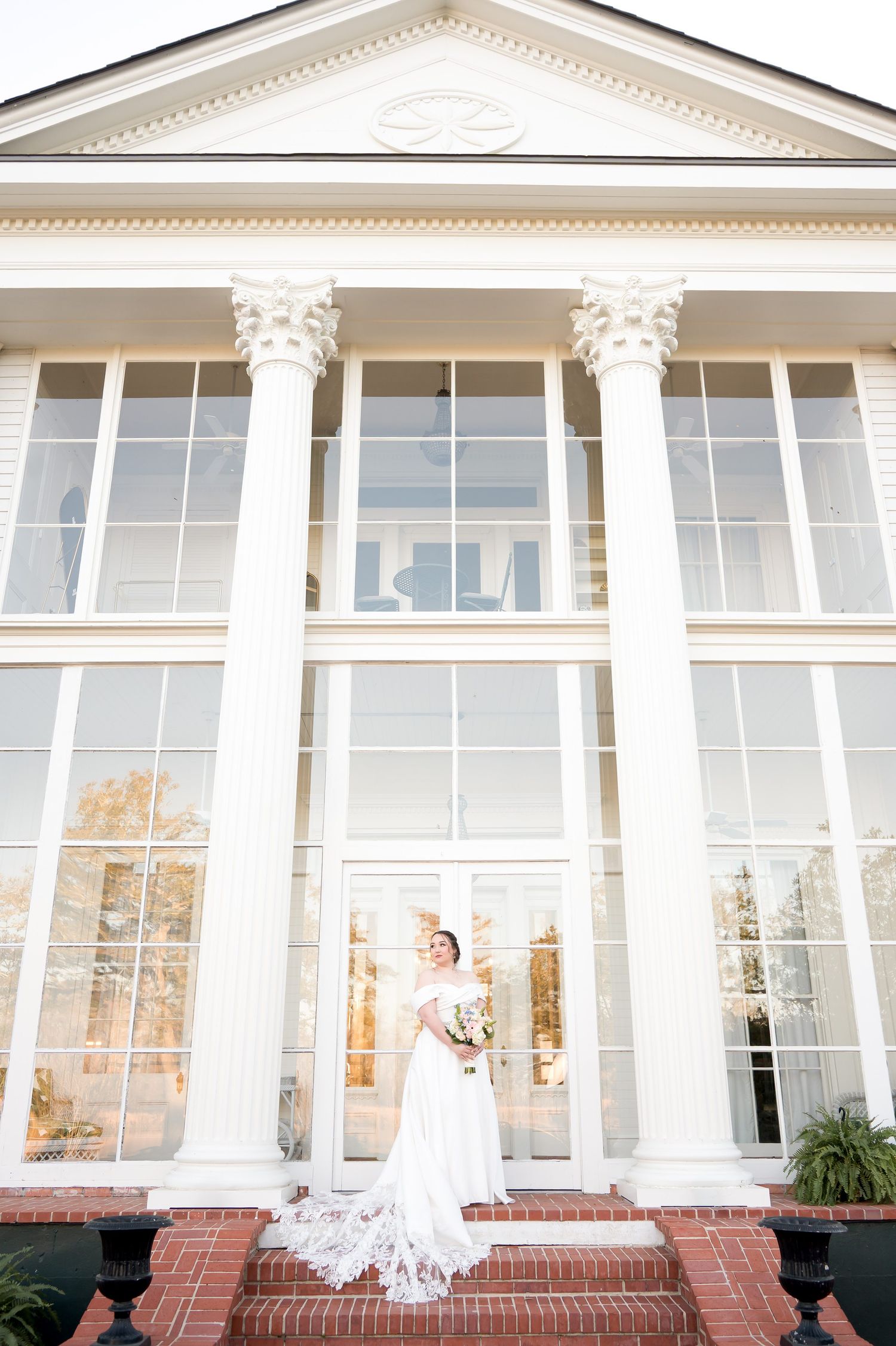 Bride in white gown stands on brick steps before grand white colonial building with tall columns and large windows.