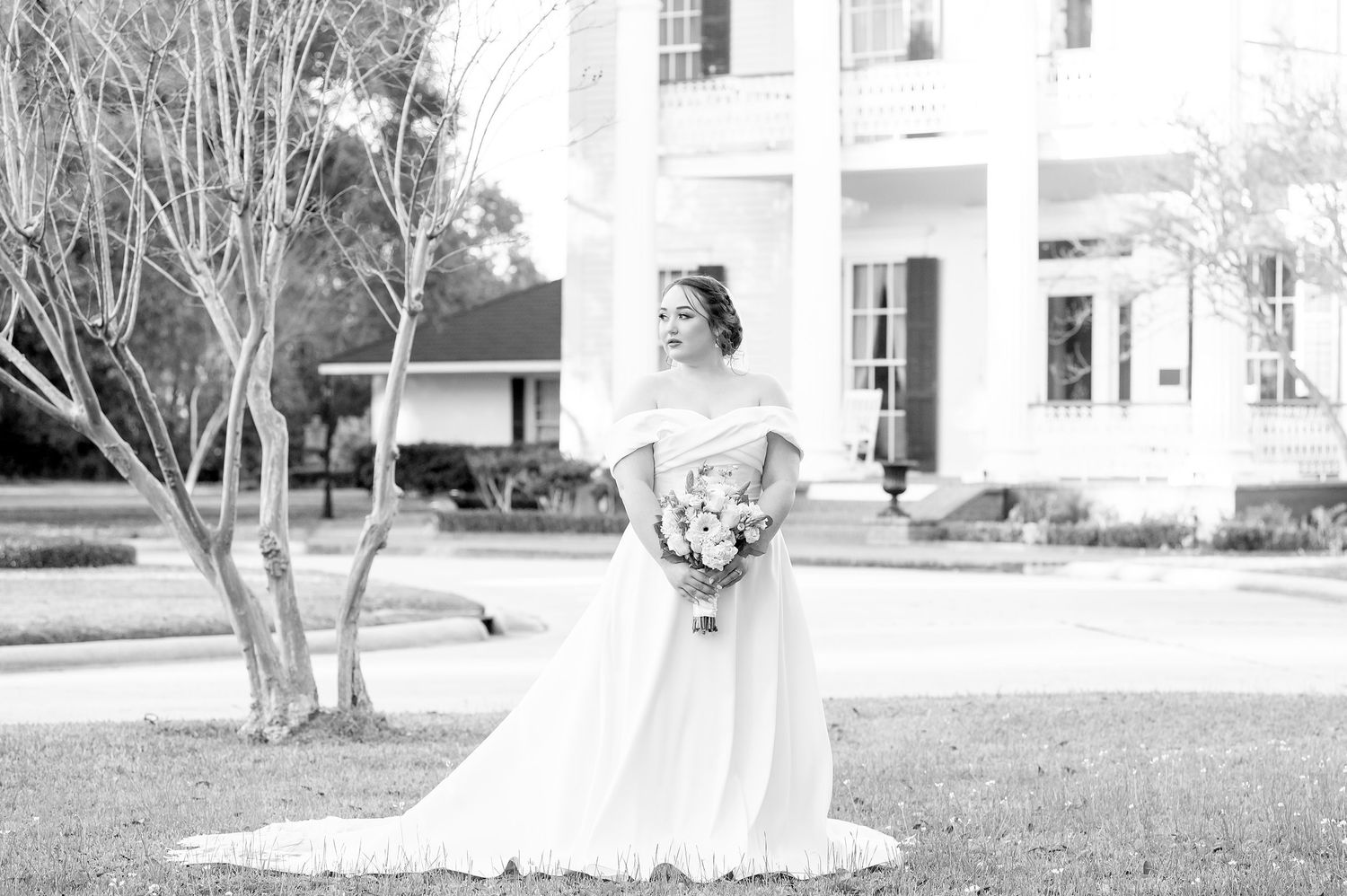 Bride in off-shoulder white gown holding bouquet poses on lawn before grand Southern mansion in black and white photo.