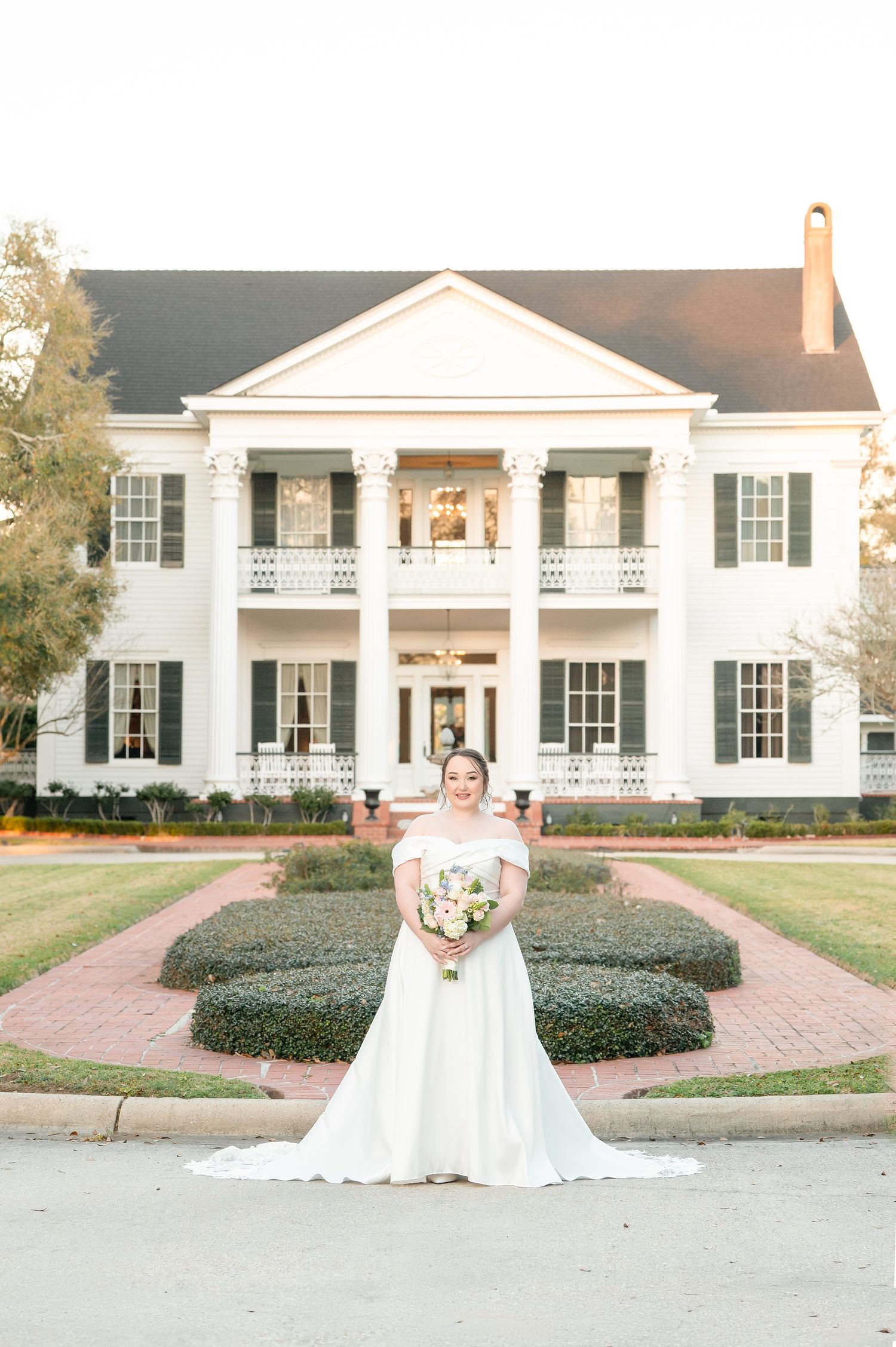 Bride in white off-shoulder gown holding bouquet stands before elegant Southern plantation-style mansion with columns.