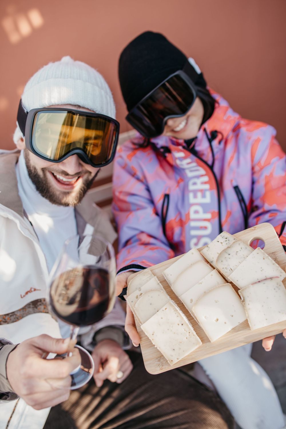 Deux skieurs en lunettes dégustent du vin et du fromage sur une planche de bois tout en se relaxant en tenue de station de ski.