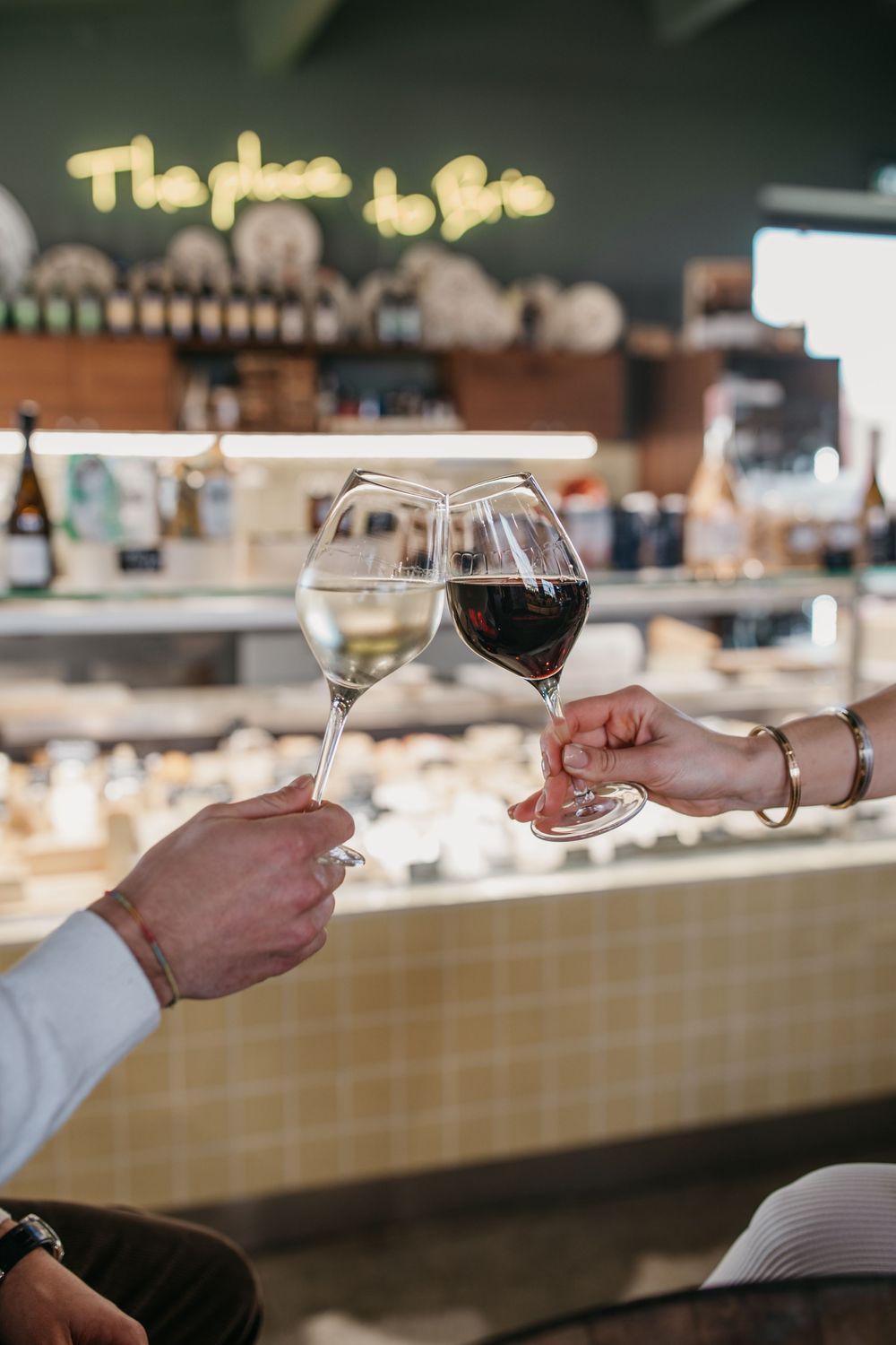 Deux mains faisant tinter des verres à vin dans un bar à fromage, avec une enseigne au néon lisant The Place to Brie en arrière-plan.