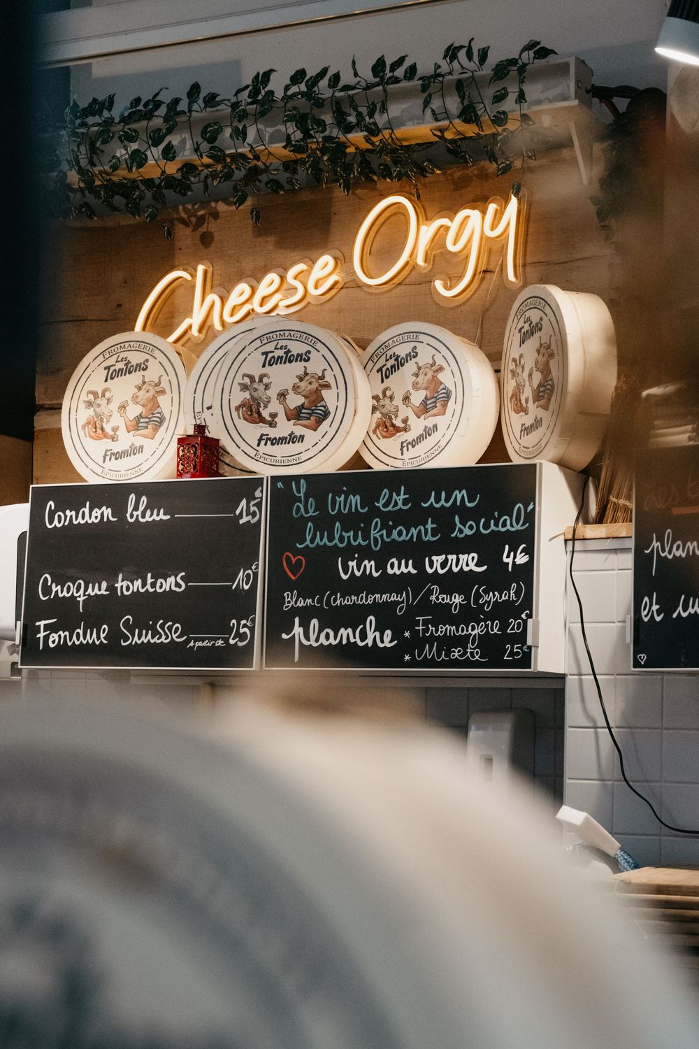 Enseigne au néon Cheese Orgy au-dessus d'un comptoir de fromagerie français avec des menus au tableau noir et des présentoirs de roue de fro