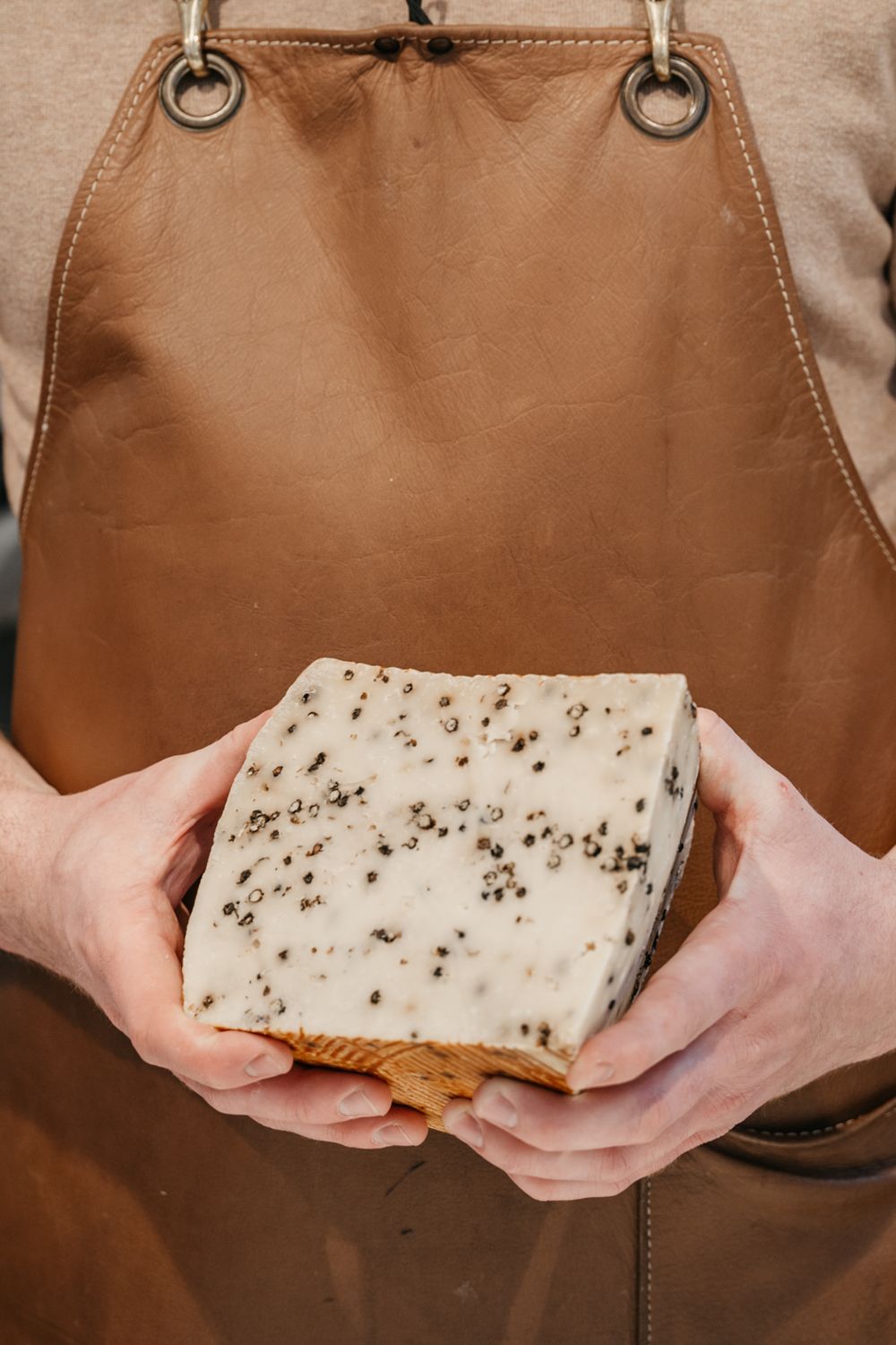 Personne en tablier en cuir marron tenant un bloc de fromage blanc parsemé de poivre à deux mains.