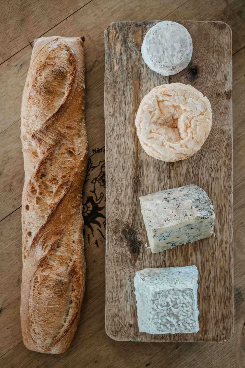 Baguette française rustique à côté d'une planche de bois avec quatre fromages artisanaux, dont du brie et du fromage bleu.