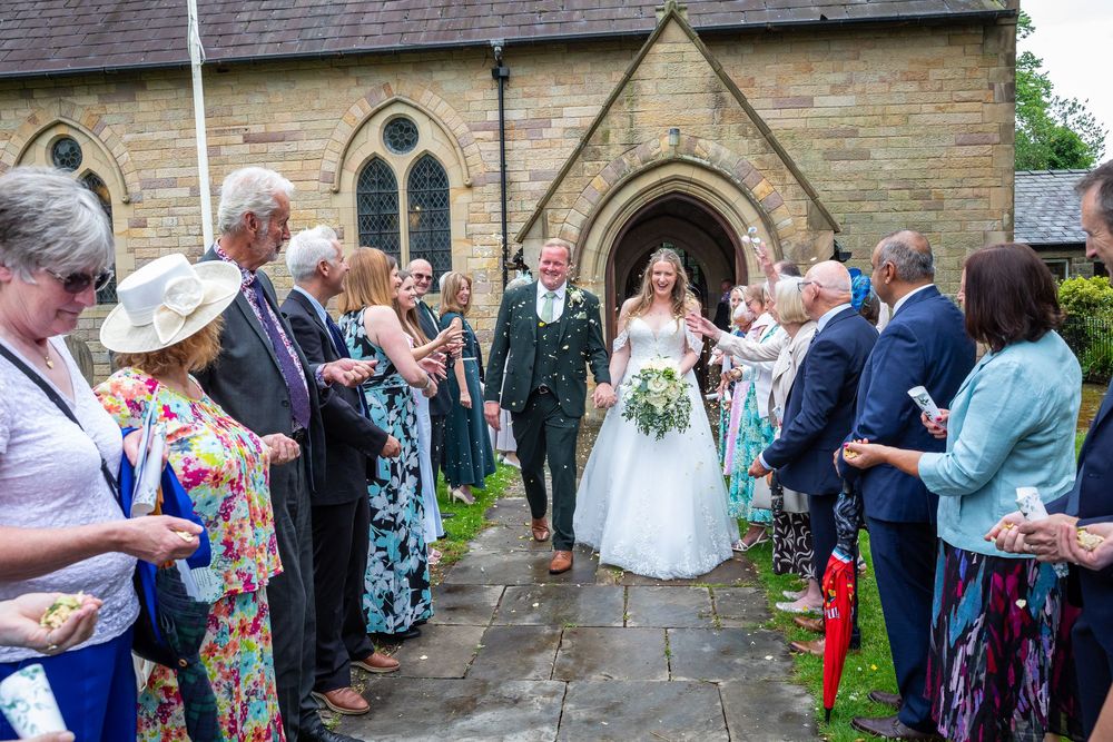 Smiling newlyweds walk through a confetti line of guests outside a traditional stone church.