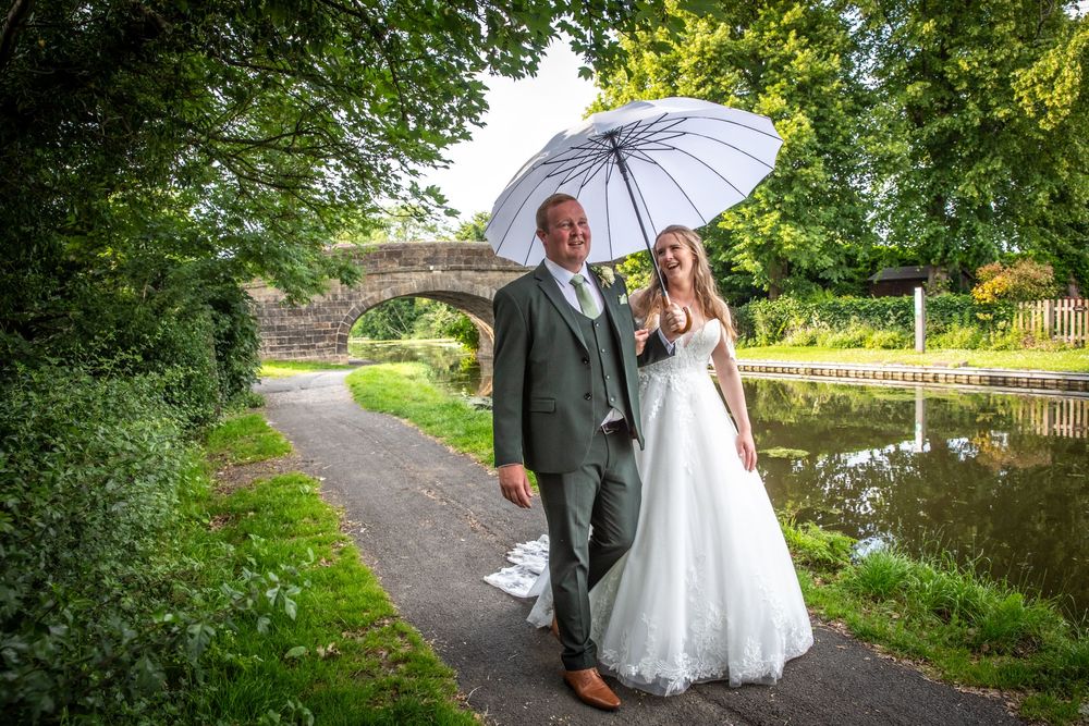 Bride and groom stroll along a tree-lined canal towpath sharing a white umbrella.