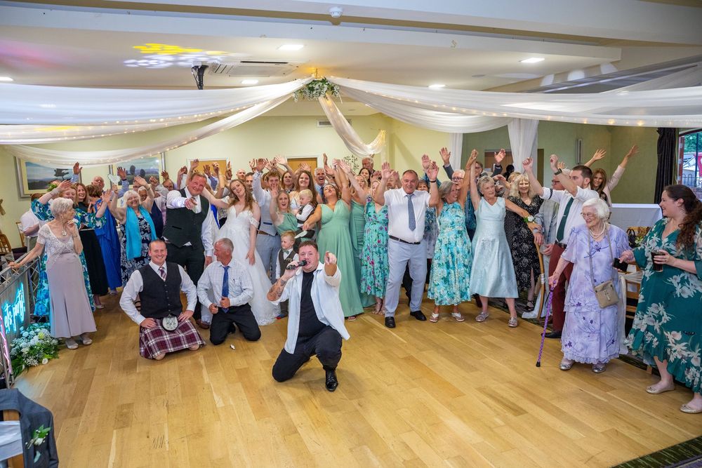 Large wedding reception group photo with guests cheering and raising hands in a decorated hall.
