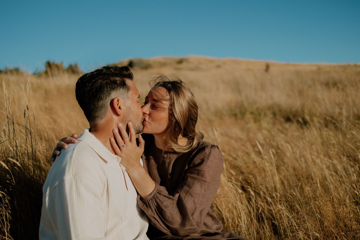 A couple shares an intimate moment in a golden wheat field against a bright blue sky during sunset.