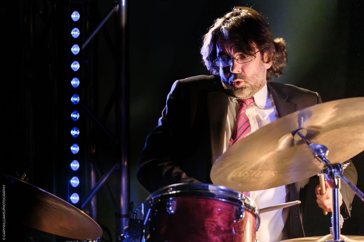 A drummer performs on stage with a drum kit in dramatic lighting against a dark background.