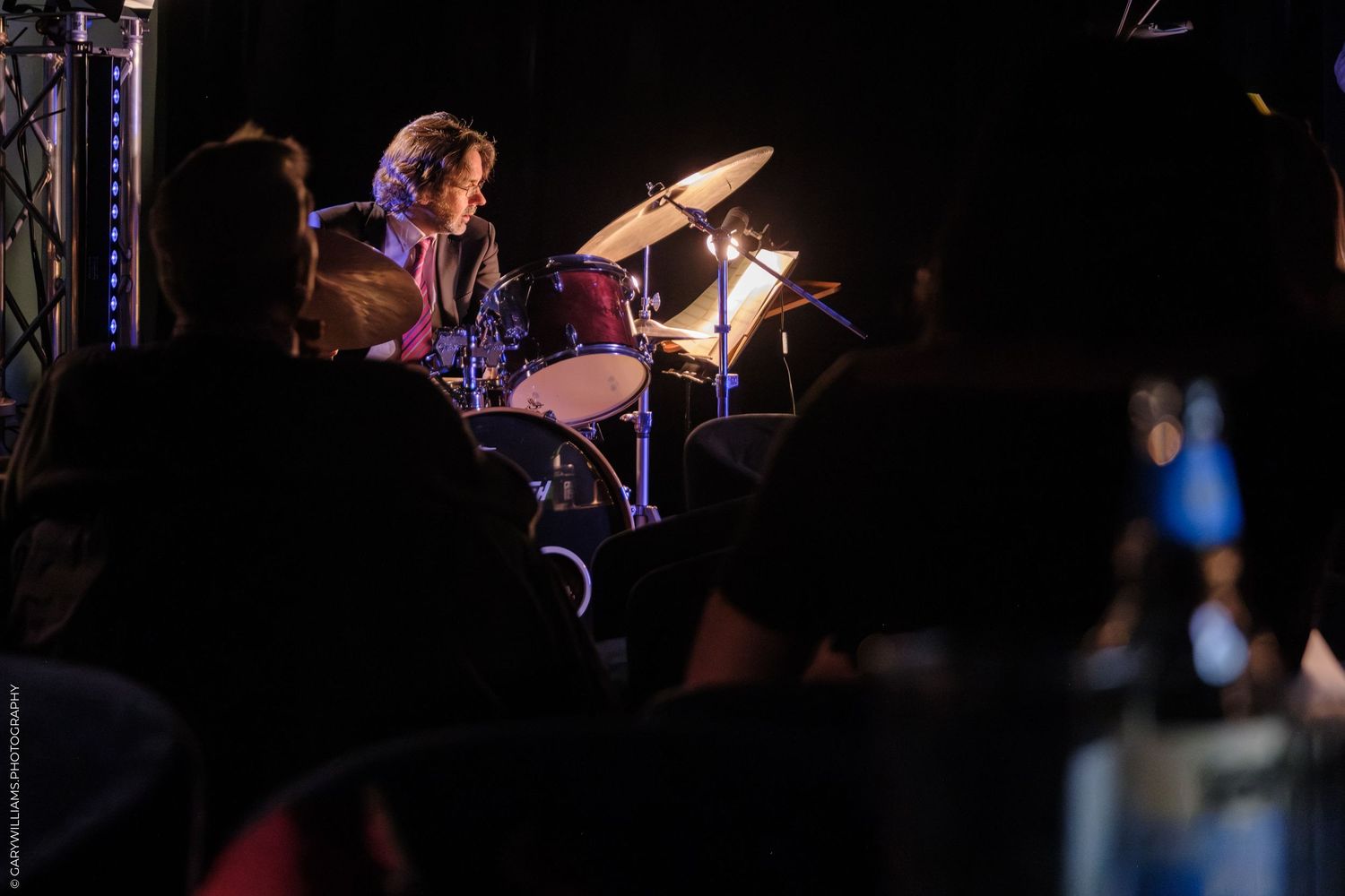 Drummer plays energetically on stage with dramatic lighting and equipment visible in background.