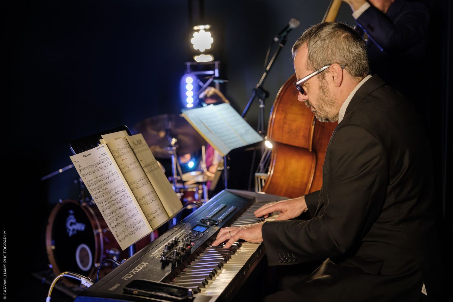 Musicians perform jazz music on stage with piano, double bass, and sheet music stands illuminated in blue lighting.