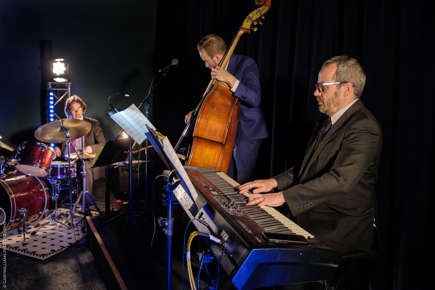 Jazz trio performs on stage with piano, upright bass and drums in dark concert lighting.
