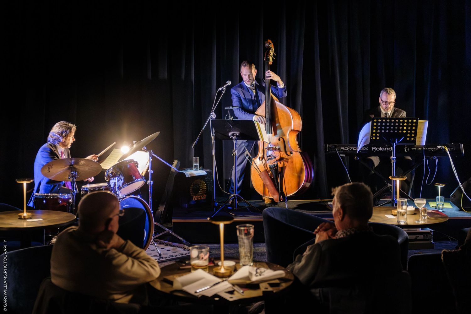 Musicians perform on stage with instruments in a dimly lit concert setting with dramatic lighting effects.