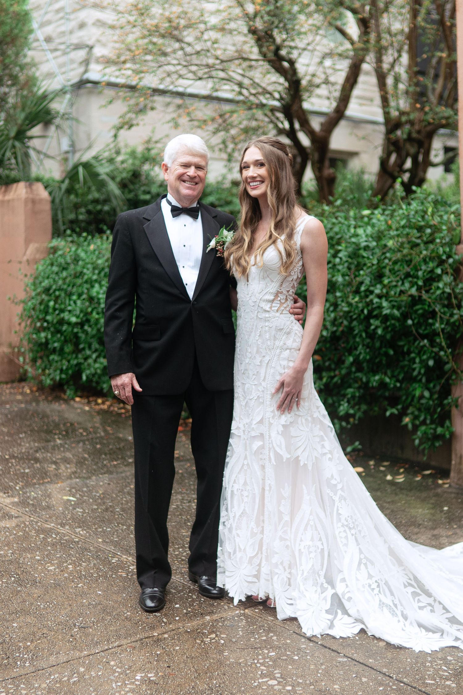 Couple in formal wedding attire posing along a garden path with lush greenery.