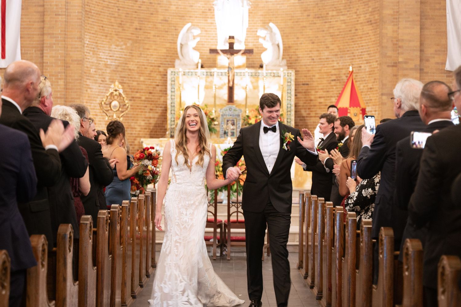 Newlyweds walk down church aisle while guests celebrate in wooden pews with ornate altar in background.