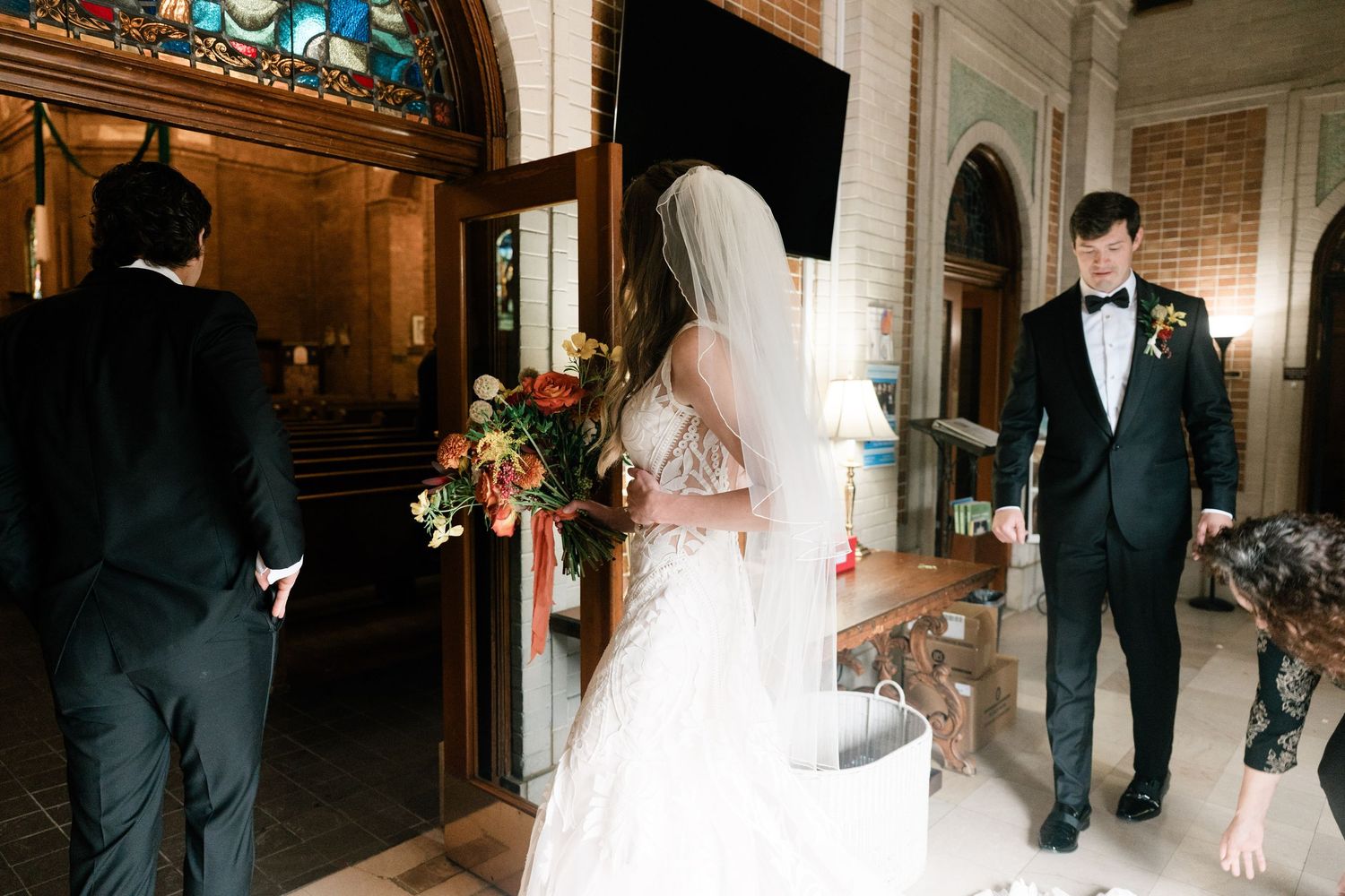A wedding party walks through an elegant hallway with ornate architectural details and warm lighting.