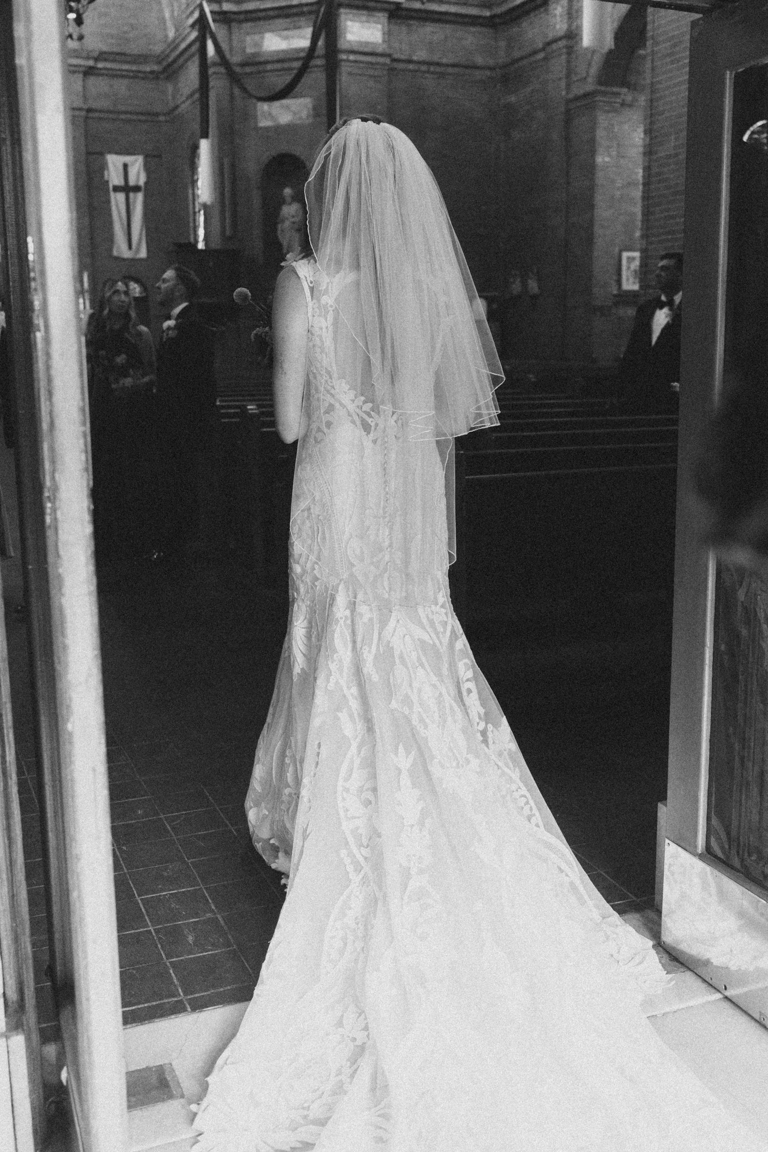 Black and white photo showing flowing wedding dress trains reflected in glass doors of church entrance.