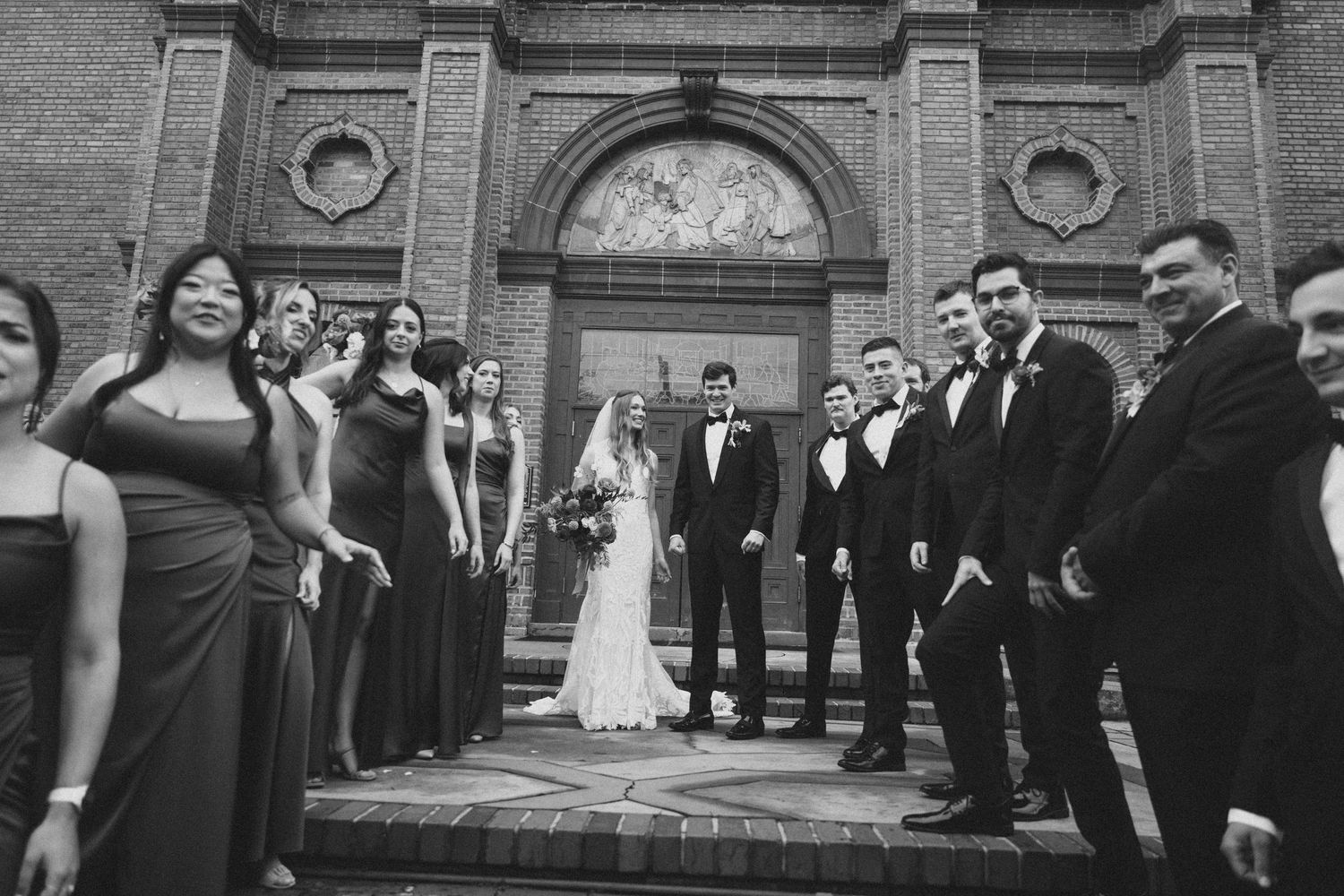 Black and white photo of wedding party celebrating on church steps with dramatic architectural backdrop.
