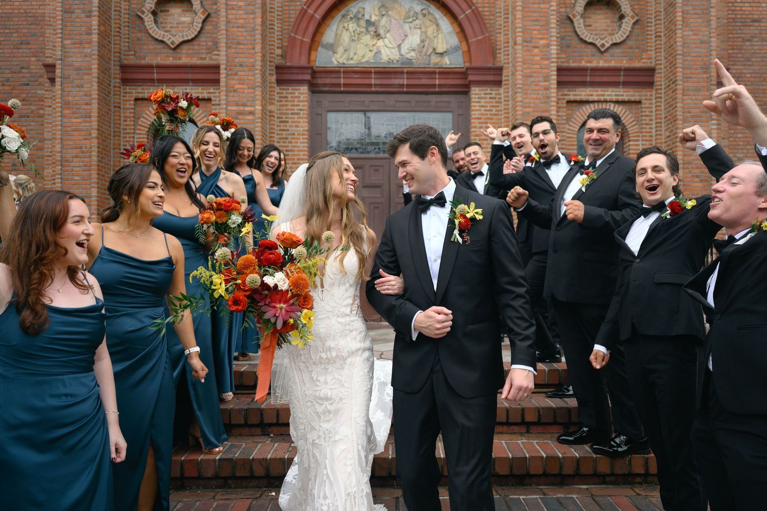 Celebratory group photo of wedding party against brick church facade with decorative details.
