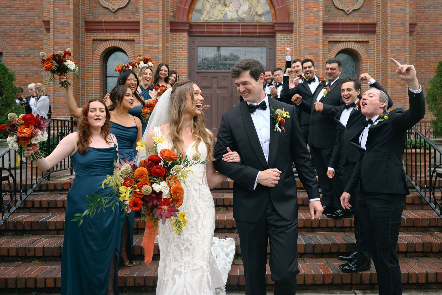 Wedding party in formal attire gathered on church steps for group photos.