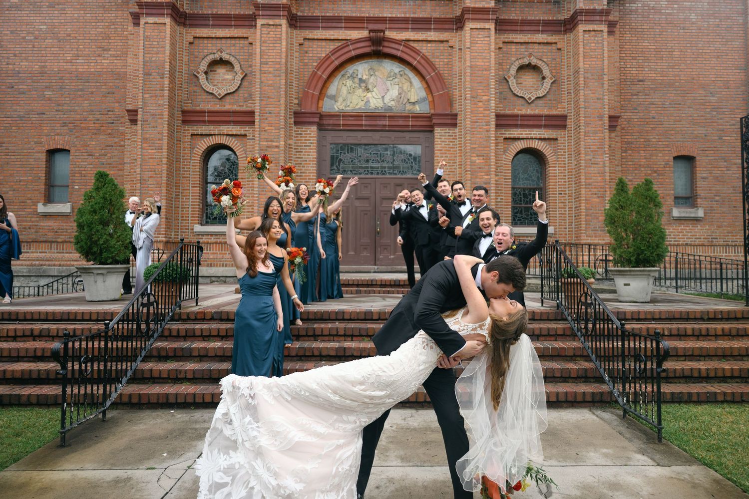 Wedding party members pose in front of a historic brick church with ornate arched doorways and iron railings.
