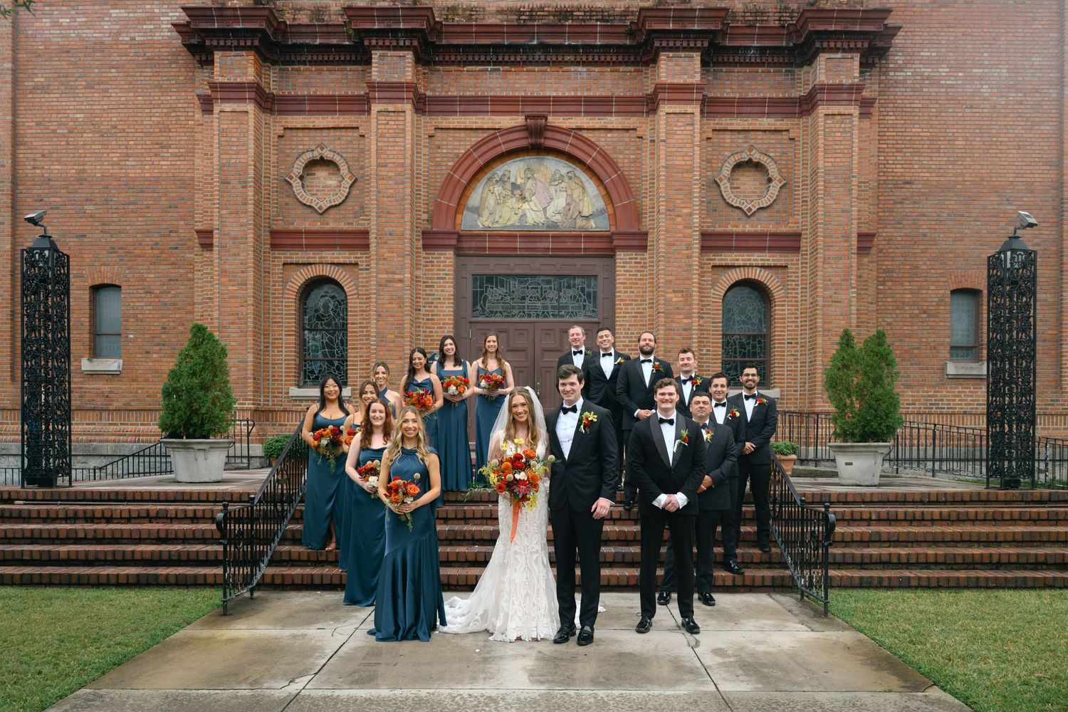 Wedding party members pose for formal photos in front of a historic brick church building with arched windows.