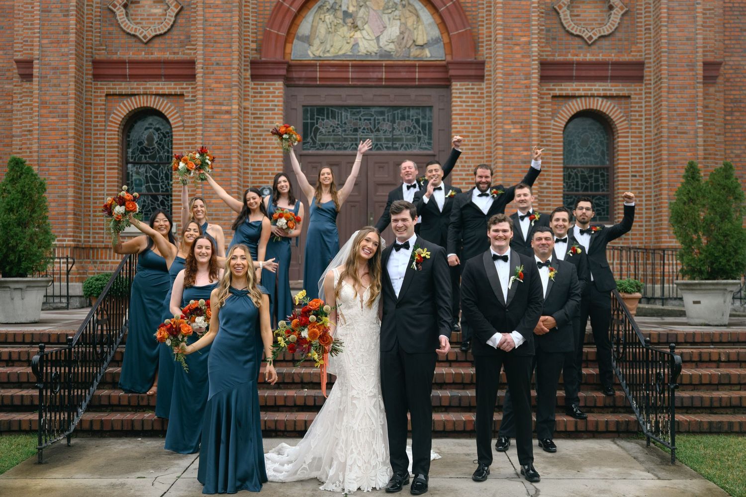 Wedding party poses joyfully on brick church steps with bridesmaids in navy dresses and groomsmen in black suits.