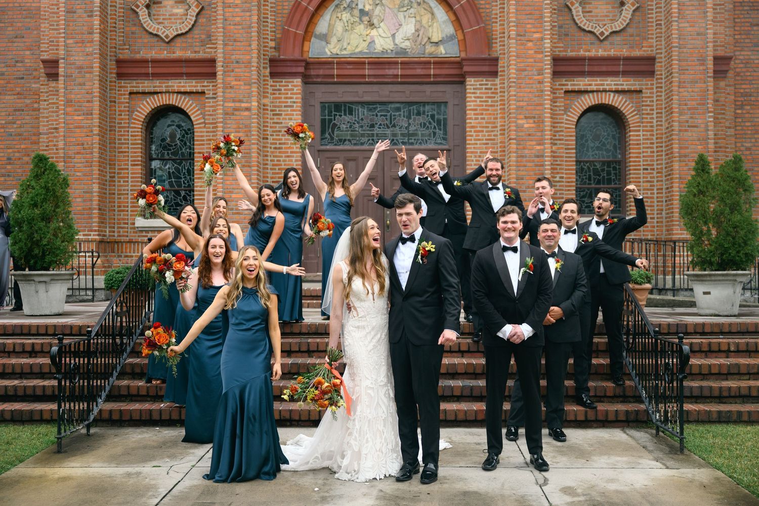 Wedding party celebrating with joyful poses outside historic brick church entrance.