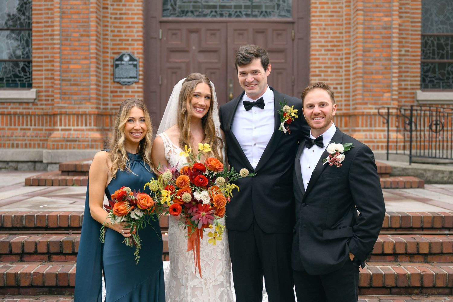 Wedding party gathers on brick steps of historic church building.