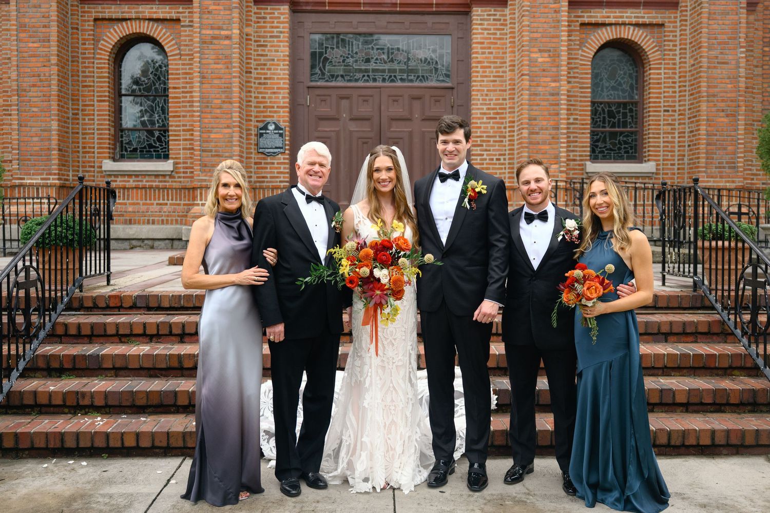 Wedding party stands on church steps with bride in white lace gown and attendants in formal attire holding autumn bouquets.