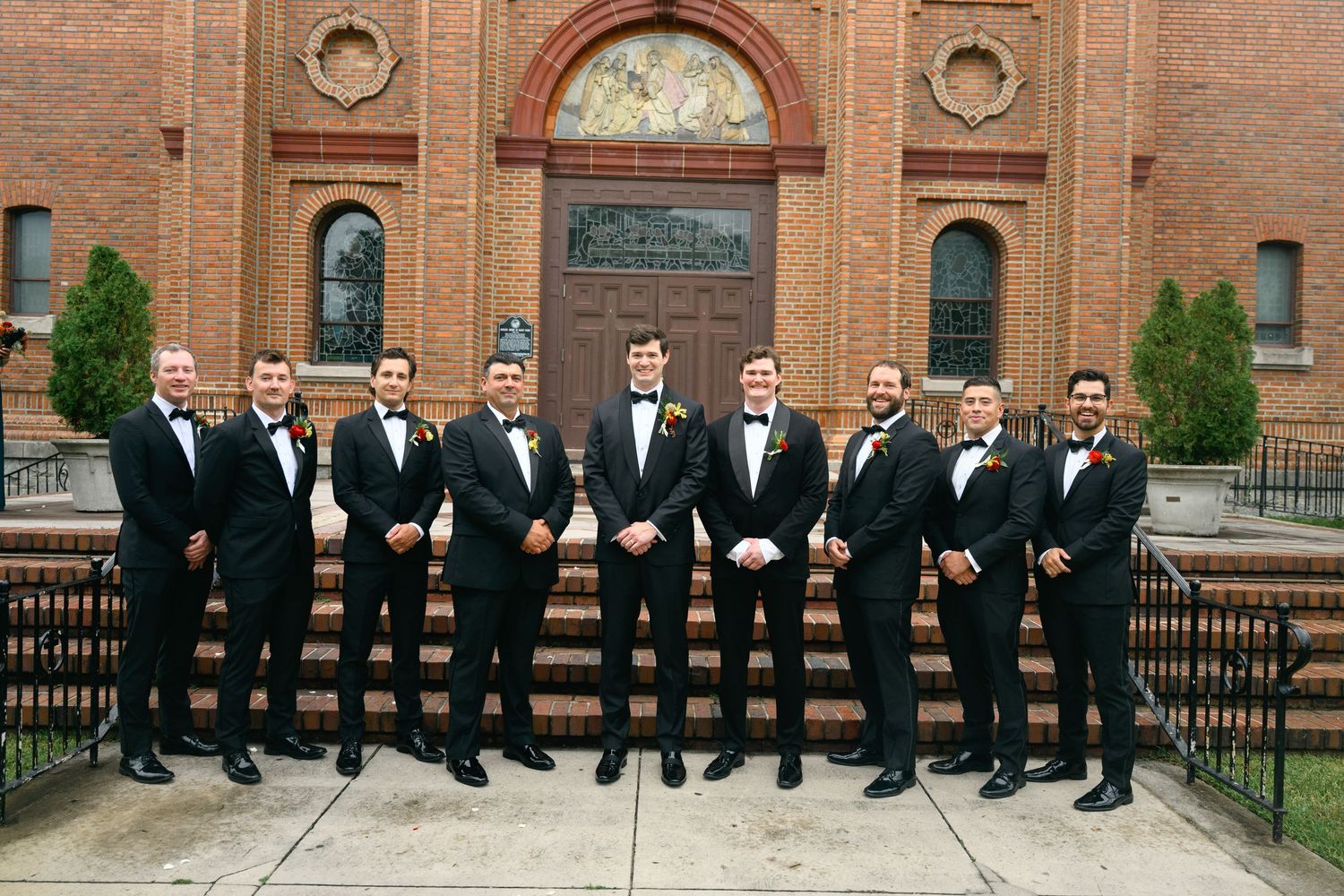 Groomsmen in black tuxedos pose together on brick church steps for formal wedding photo.