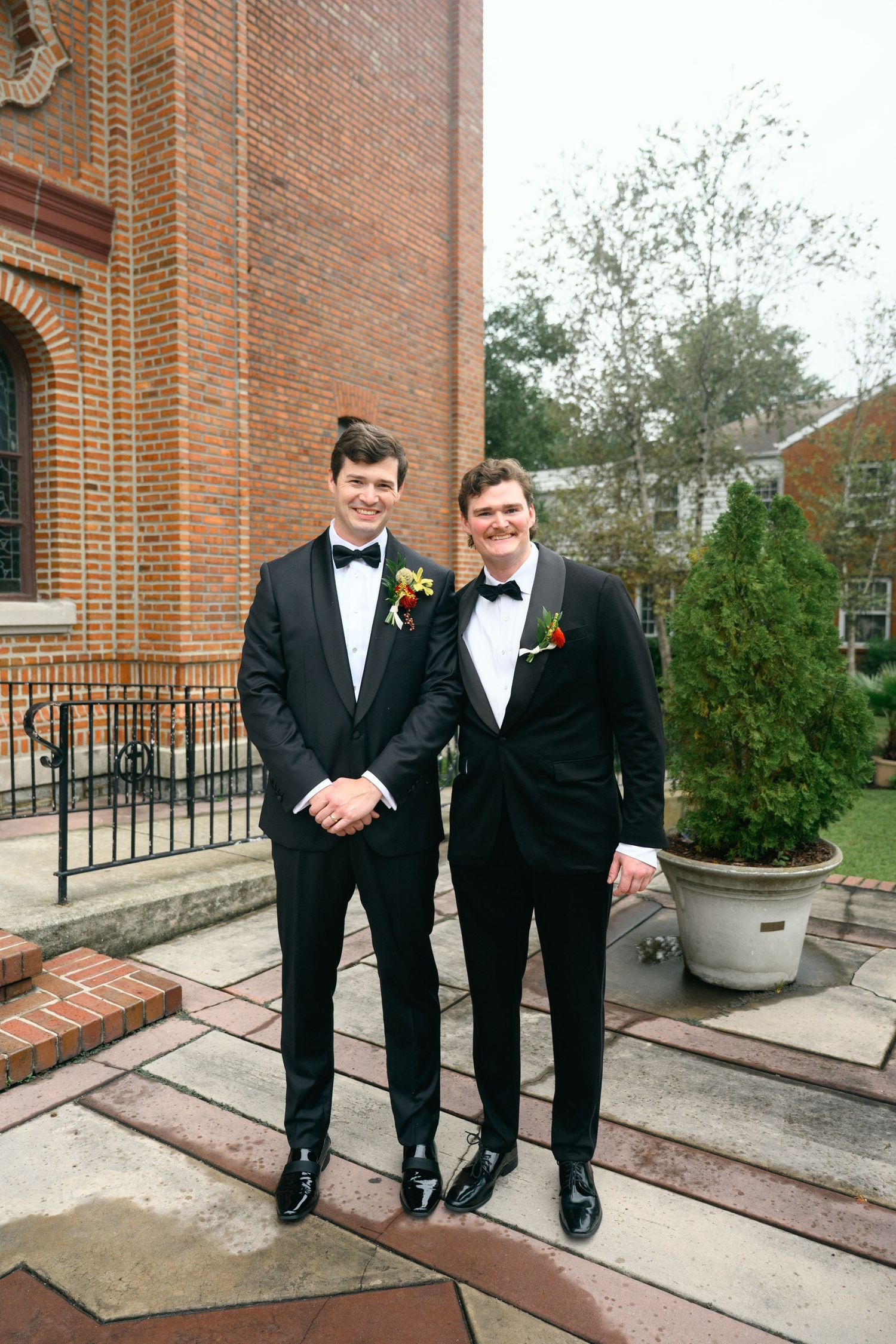 Groomsmen in black tuxedos and bowties pose together outside a brick church building.