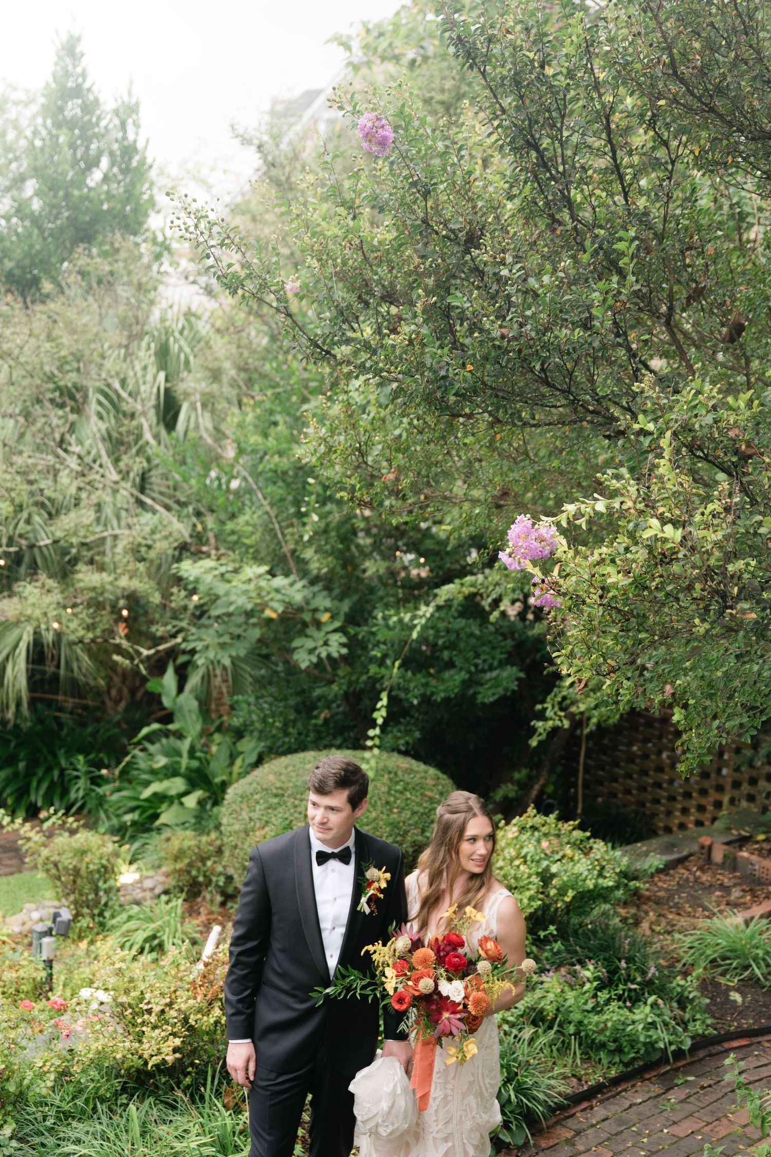 Wedding couple poses in lush garden setting with vibrant fall-colored bouquet.