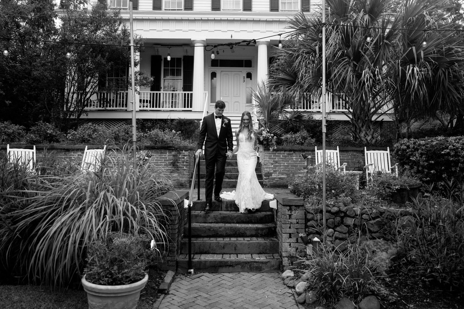 A couple stands on the steps of a grand white colonial house with a wraparound porch and lush garden.