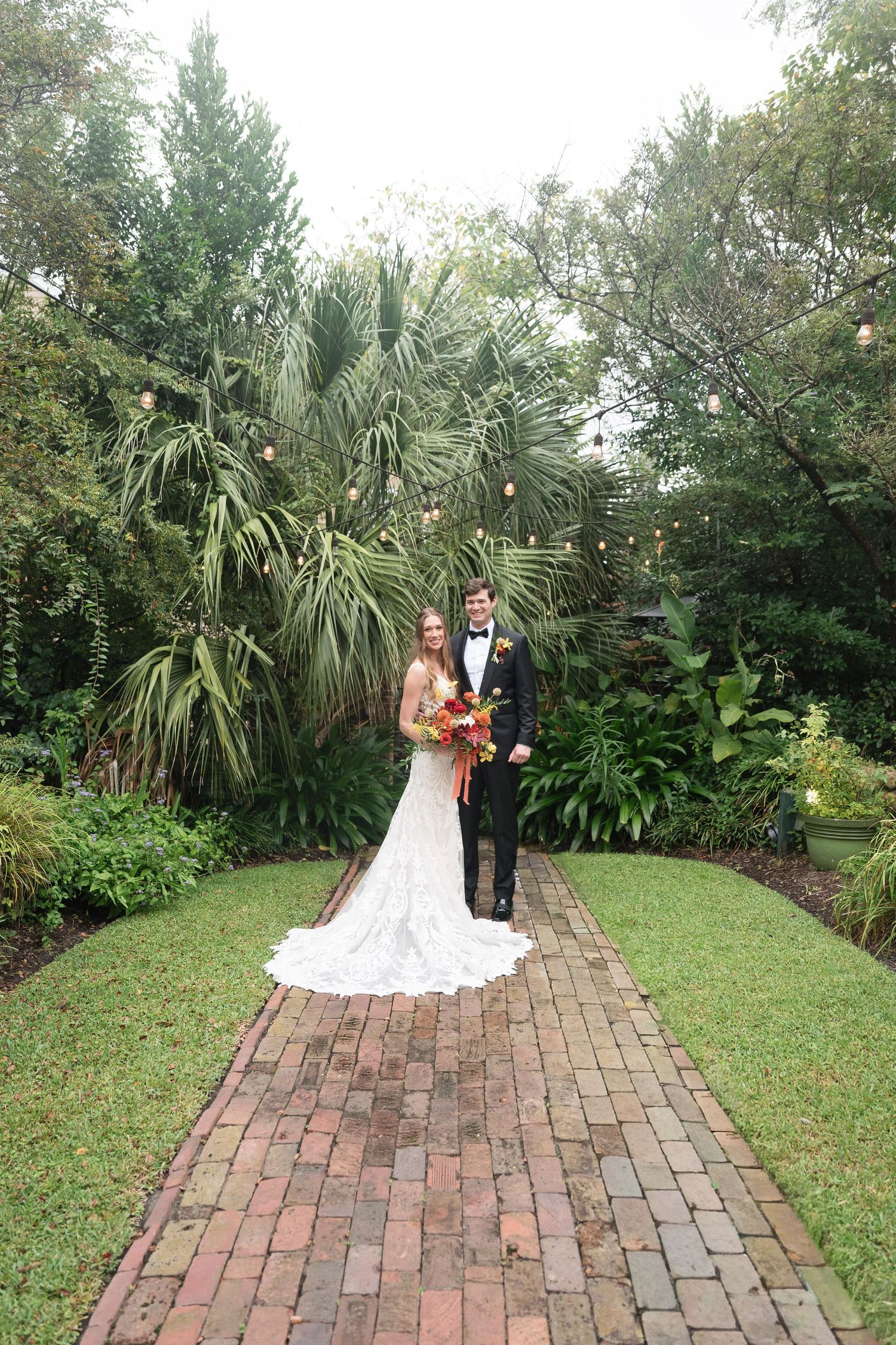 A couple poses on a brick pathway surrounded by lush tropical gardens and palm trees.