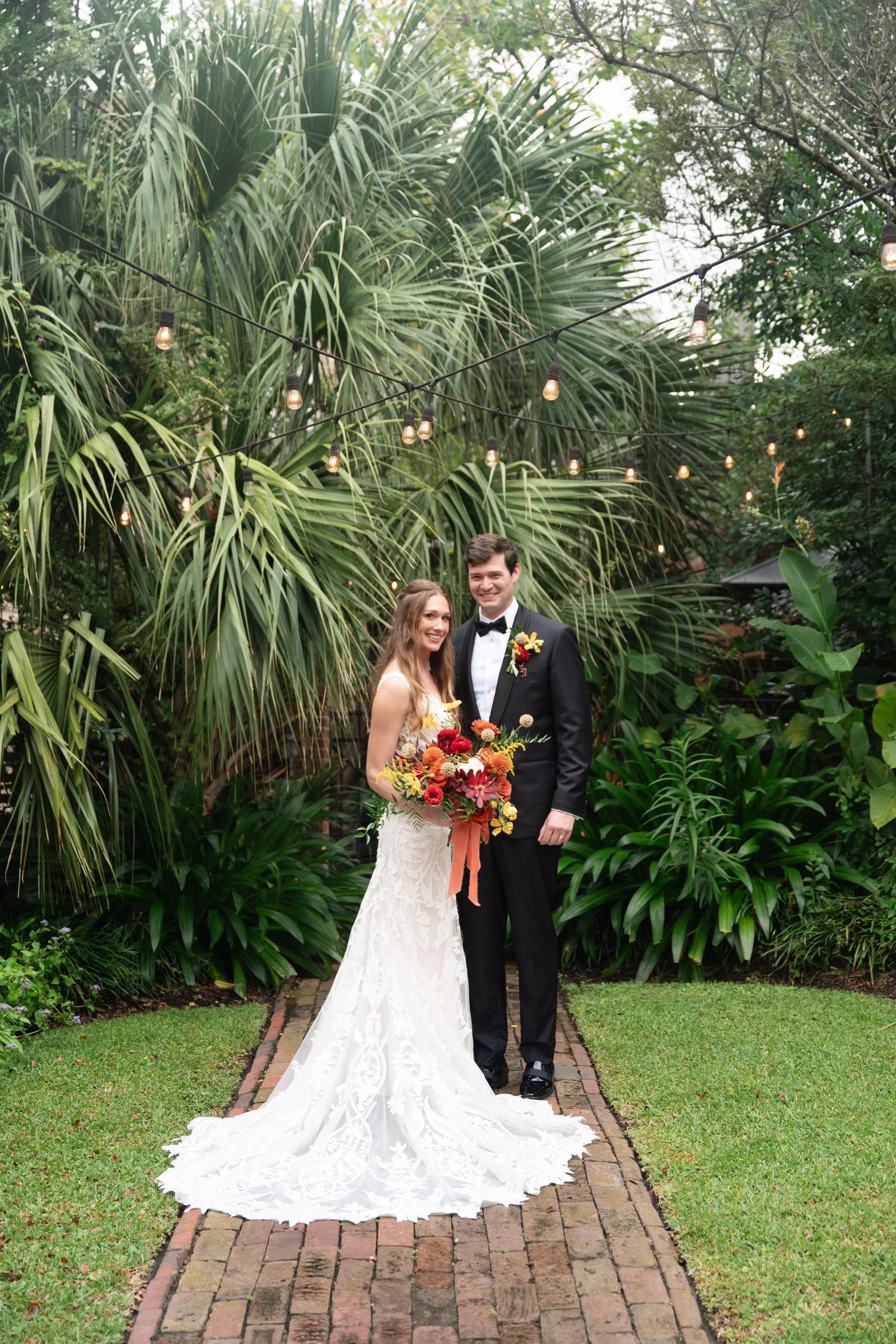 A couple in wedding attire poses on a brick pathway surrounded by lush tropical palm trees and greenery.