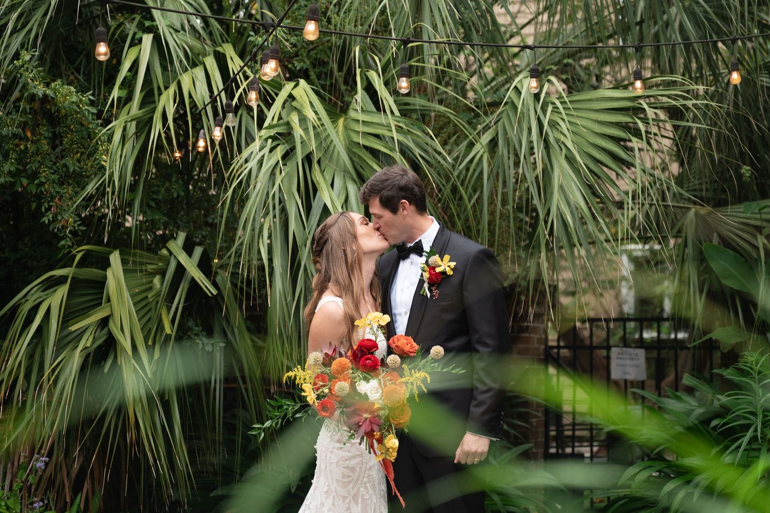 Newlyweds embrace in a garden setting with palm trees and string lights creating a magical atmosphere.