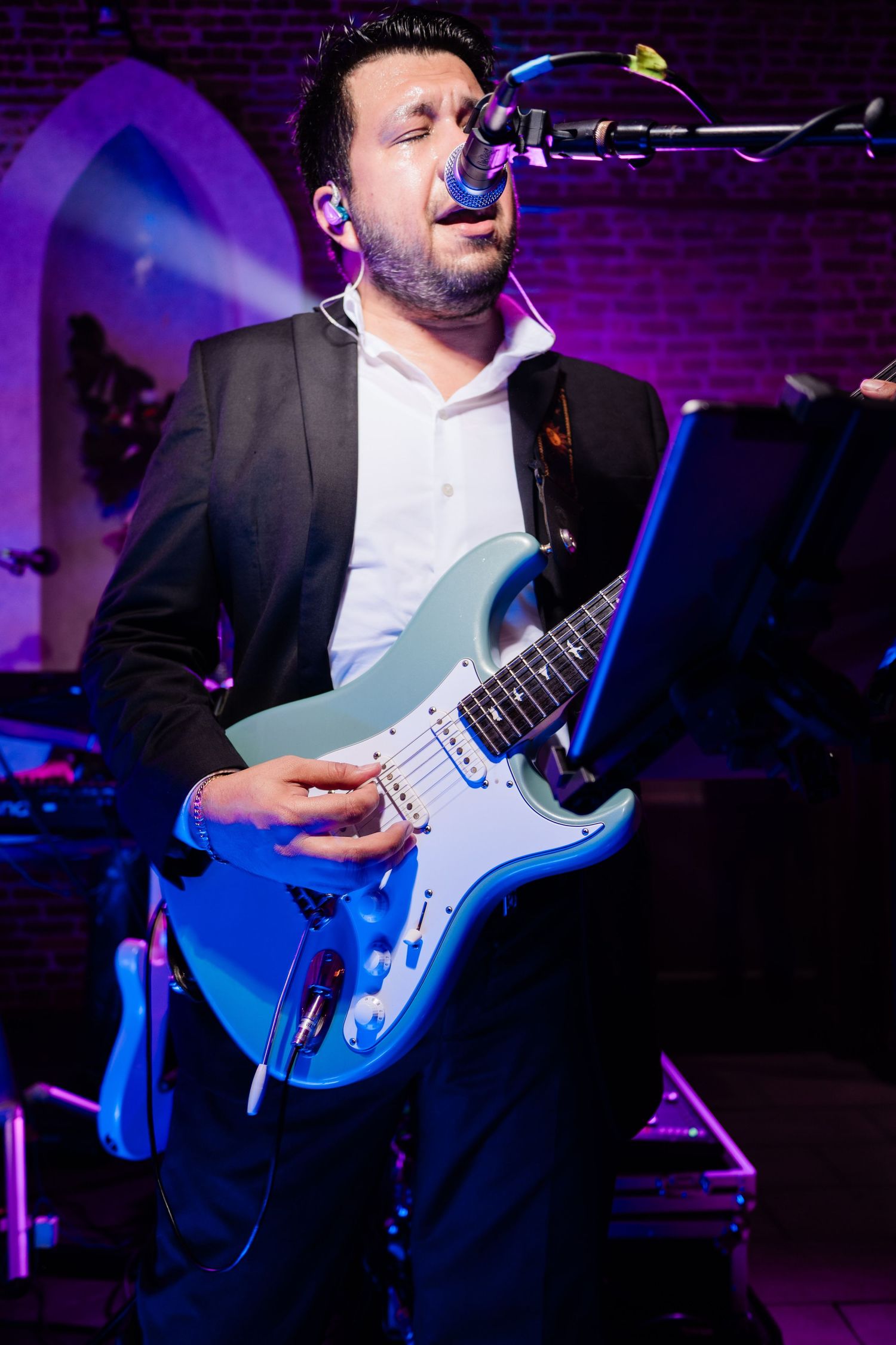 Musician playing electric blue guitar under dramatic purple stage lighting at wedding reception.
